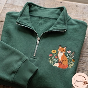 Könnte beinhalten: Ein dunkelgrünes Sweatshirt mit kurzem Reißverschluss und einer Stickerei, die einen Fuchs inmitten bunter Blumen zeigt. Das Kleidungsstück verfügt über einen silberfarbenen Metallreißverschluss, einen Stehkragen und gerippte Bündchen.
