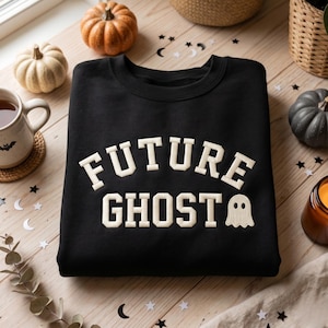 May include: A black crewneck sweatshirt featuring the text FUTURE GHOST in a cream-colored collegiate font, with a small ghost graphic positioned to the right of the word GHOST. The design is centered on the front of the garment.