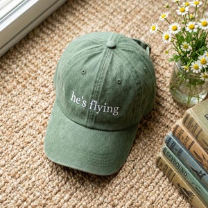 Puede incluir: Gorra de béisbol verde salvia con el texto bordado "he's flying" en blanco. La gorra está hecha de un material suave y tiene una visera curva. La gorra está sobre una superficie tejida.