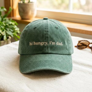 Puede incluir: Una gorra de béisbol de color verde bosque con una corona estructurada y una visera curva. La parte frontal de la gorra presenta el texto bordado "hi hungry, i'm dad." en una fuente sencilla y clara, centrado sobre la visera.