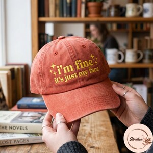 May include: A rust-colored baseball cap with a distressed finish featuring the yellow embroidered text "I'm fine it's just my face" accompanied by small star icons. The cap has a curved brim and a button on the top center of the crown.