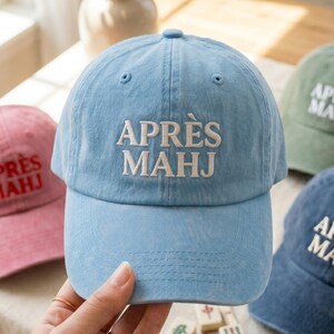 May include: Light blue baseball cap with the embroidered text "APR&Egrave;S MAHJ" in white. The cap has a curved brim and a rounded crown. Other caps in pink, green, and blue are visible in the background.