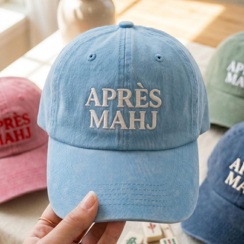 May include: Light blue baseball cap with the embroidered text "APR&Egrave;S MAHJ" in white. The cap has a curved brim and a rounded crown. Other caps in pink, green, and blue are visible in the background.