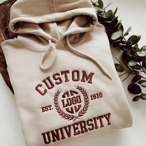 Może przedstawiać: Beżowa bluza z kapturem z ciemnobrązowym haftem o treści CUSTOM UNIVERSITY oraz napisem EST. 1910, który otacza centralny okrągły emblemat z wyhaftowanym słowem LOGO wewnątrz wieńca laurowego.
