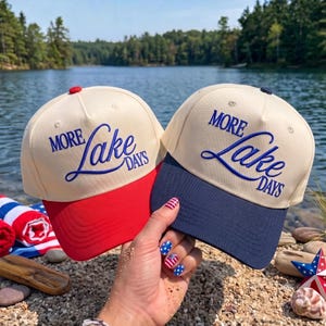 Könnte beinhalten: Zwei Baseballkappen mit cremefarbenen Kronen und blauem Sticktext, der MORE Lake DAYS lautet. Eine Kappe hat einen einfarbig roten Schirm und einen roten Knopf oben, während die andere Kappe einen einfarbig marineblauen Schirm besitzt.
