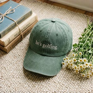 May include: A green baseball cap with the text "he's golfing" in white. The hat is on a woven rug next to a stack of books and a bouquet of white flowers with yellow centers. The books are titled "Golfing Memoirs", "The Links Journey", and "A History of the Green".