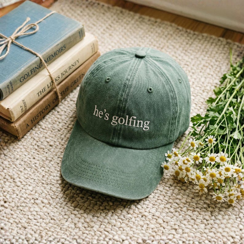 May include: A green baseball cap with the text "he's golfing" in white. The hat is on a woven rug next to a stack of books and a bouquet of white flowers with yellow centers. The books are titled "Golfing Memoirs", "The Links Journey", and "A History of the Green".