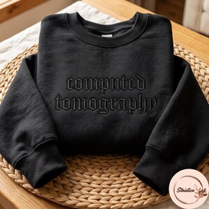 May include: A black crewneck sweatshirt featuring the words computed tomography embroidered in a dark, tonal gothic-style font across the chest. The garment has long sleeves with ribbed cuffs and a ribbed neckline, made of a solid black fabric.