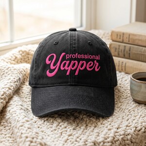 May include: A black baseball cap with the words "professional Yapper" embroidered in pink. The hat is on a cream-coloured knitted blanket, with books and a mug in the background.