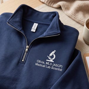 May include: A navy blue quarter-zip sweatshirt featuring a white embroidered microscope icon and the text "Olivia, MLS (ASCP) Medical Lab Scientist" on the left chest. The garment has a silver-toned metal zipper closure at the collar.