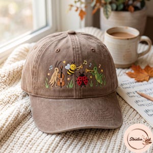 May include: A brown baseball cap featuring an embroidered design of a cricket playing a cello, a bee playing a trumpet, a ladybug, and a grasshopper playing a trumpet, surrounded by small musical notes, stars, and green grass blades.