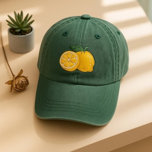 May include: A dark green baseball cap with a curved brim. The cap features an embroidered design of two yellow lemons with a green leaf. The cap is made of a soft fabric and has a button on top.
