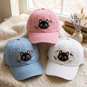 May include: Three baseball caps in pink, light blue, and white, each featuring an embroidered black cat face surrounded by small fish, fish skeletons, and star motifs. The caps have a structured crown, a curved brim, and stitched eyelets.