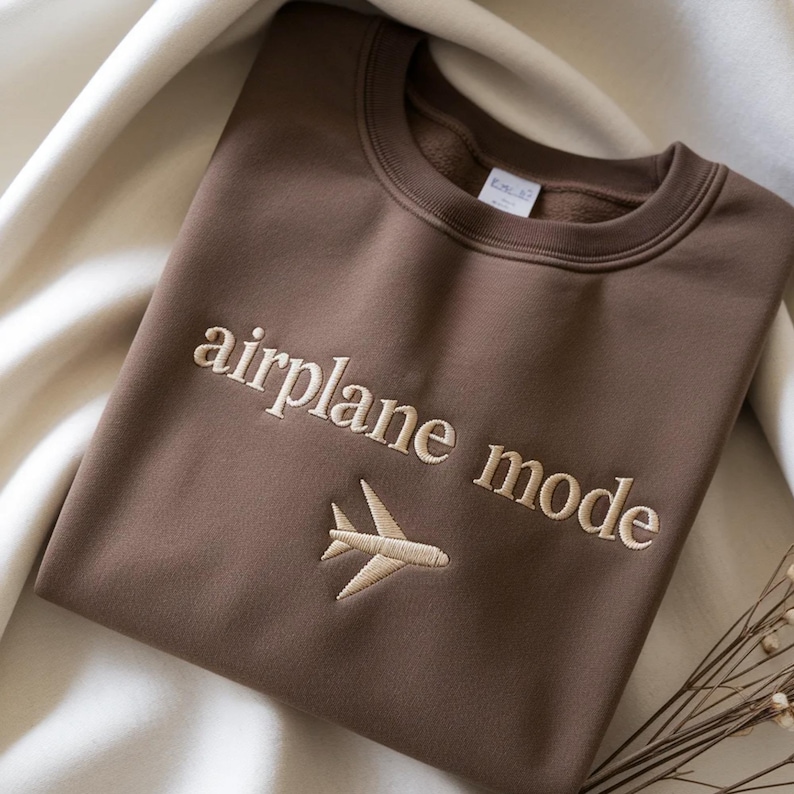 May include: A brown sweatshirt with the words "airplane mode" embroidered in a light beige color, along with a small airplane graphic. The sweatshirt is folded on a white surface.