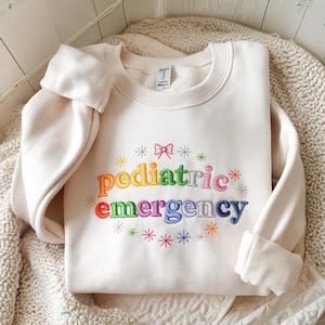 May include: Cream-colored sweatshirt with the words "pediatric emergency" embroidered in rainbow colors. The design includes snowflakes and a bow. The sleeves are cuffed.