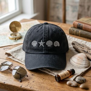 May include: A dark gray baseball cap with embroidered white seashells and a starfish. The cap is sitting on a wooden table with sunglasses, a compass, and books. The hat has a curved brim.