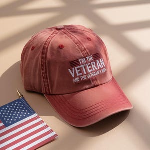 May include: A maroon baseball cap with the text "I'M THE VETERAN AND THE VETERAN'S WIFE" in white. The cap has a curved brim and a button on top. An American flag is in the lower left corner.