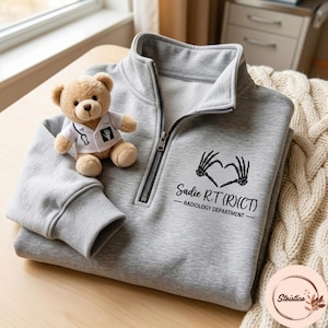 May include: A light gray quarter-zip sweatshirt featuring a black graphic of skeleton hands forming a heart shape above the text Sadie R.T (R)(CT) RADIOLOGY DEPARTMENT. A small tan teddy bear wearing a white lab coat with a stethoscope is included.