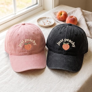 May include: Two baseball caps, one in dusty pink and one in charcoal black, each featuring the embroidered text "just peachy" in a cream-colored font arched above a small, stylized peach graphic with green leaves centered on the front panel.