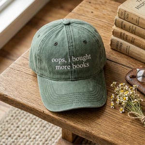 May include: A green baseball cap with the text "oops, i bought more books" in white. The cap is sitting on a wooden table next to a stack of books and a small bouquet of flowers.