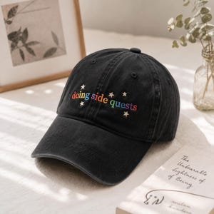 May include: A black baseball cap featuring the embroidered text "doing side quests" in a rainbow gradient font. The design includes small embroidered stars surrounding the text. The cap has a structured crown and a curved brim.