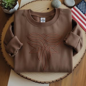Pu&ograve; includere: Una felpa marrone con girocollo e maniche lunghe. La felpa presenta un design astratto ricamato in una tonalit&agrave; pi&ugrave; chiara di marrone sul davanti. L'indumento &egrave; esposto su una superficie di legno, con una pianta succulenta e una bandiera sullo sfondo.