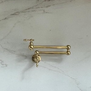 May include: A brass wall-mounted pot filler faucet with a double-jointed swing arm, positioned above a black gas stovetop with a central griddle. To the left, two wooden cutting boards lean against a white marble backsplash with grey veining.