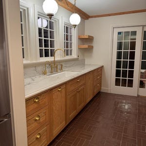 May include: A kitchen interior featuring light oak wood cabinets with brass knobs, a white marble countertop with a white sink, and a gold faucet. Two white globe pendant lights hang above the counter, and the floor is made of brown brick tiles.