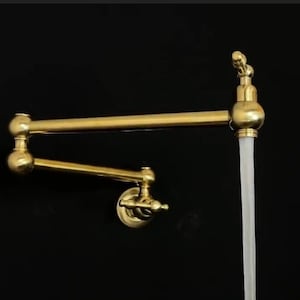 Op de afbeelding: Een gouden keukenkraan met een zwenkbare uitloop en een lange, dunne straal water die eruit stroomt. De kraan is aan een muur gemonteerd en heeft een decoratief ontwerp.