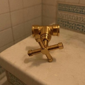May include: A polished gold-toned metal faucet handle featuring a classic cross-shaped design with four extending arms. The fixture has a central hub and a cylindrical stem, showcasing a metallic finish that reflects light across its smooth surfaces.
