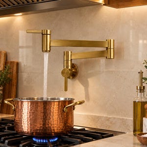 Op de afbeelding: Een aan de muur gemonteerde, geborsteld gouden pannenvul-kraan met een dubbel scharnierende zwenkarm, die een constante waterstraal in een grote, ronde, gehamerde koperen pan met twee zijhandgrepen giet, staand op een zwarte gasfornuisbrander.