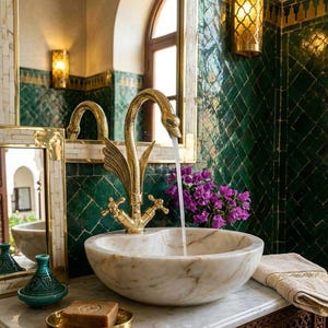 May include: A gold swan-shaped faucet pours water into a round white marble vessel sink. The setup includes a small teal ceramic lidded jar, a bar of soap on a gold dish, and a beige towel. The background features dark green diamond-patterned tiles.