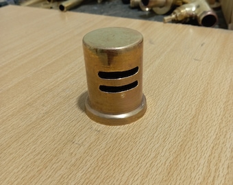May include: A gold-colored metal pipe fitting with two slots on the side. The fitting is on a light brown wooden surface.