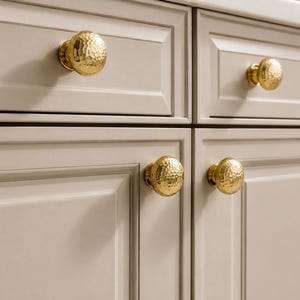 Puede incluir: Cuatro pomos redondos para armarios con un acabado dorado pulido y una textura martillada distintiva. Cada pomo está montado en una puerta de armario o frente de cajón de color beige claro con un diseño de panel empotrado y bordes rectos.