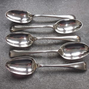 May include: Five silver spoons on a grey surface. The spoons have oval bowls and long, slender handles. The spoons are highly reflective, with the environment mirrored in their polished surfaces.