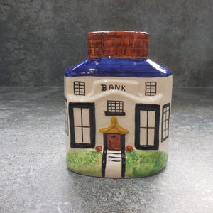 Könnte beinhalten: Eine Keramik-Spardose in Form eines Gebäudes, mit blauem Dach und ziegelartiger Oberseite. Das Wort "BANK" steht über den Fenstern. Das Gebäude hat schwarze Fensterrahmen, eine Tür und grünes Gras am Sockel.