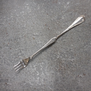 May include: A silver fork with a long handle and three tines. The handle has a decorative pattern near the top and a series of small dots in the middle. The fork is likely for serving or eating.