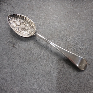 Pode incluir: Uma colher de prata com uma tigela ornamentada e detalhada com um desenho floral e de uvas. A colher tem um cabo longo e fino e está colocada sobre um fundo cinzento. A colher é provavelmente vintage ou antiga.