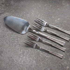 Op de afbeelding: Een roestvrijstalen taartschep en vier bijpassende vorken zijn gerangschikt op een grijze ondergrond. De schep heeft een breed, ovaal blad. De vorken hebben een decoratief patroon op de handvatten.