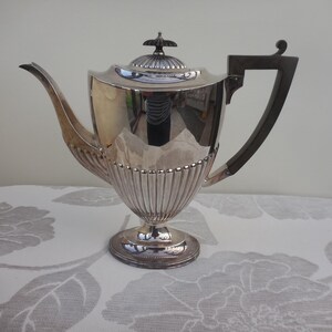 Puede incluir: Una tetera plateada con un asa oscura y una tapa decorativa. La tetera tiene un cuerpo acanalado y se asienta sobre una base redonda. El asa es oscura y angular, contrastando con la superficie plateada brillante.