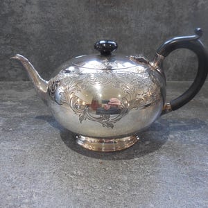 Peut inclure: Une théière argentée avec des gravures florales. Elle a une poignée noire et un bouton noir sur le couvercle. La théière est posée sur une surface grise.