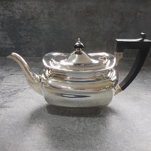 Op de afbeelding: Een gepolijste zilveren theepot met een rechthoekig lichaam met afgeronde hoeken, een gebogen tuit en een zwart handvat. Het deksel is voorzien van een kleine zwarte knop en het ontwerp heeft een reflecterende metalen afwerking met strakke lijnen.