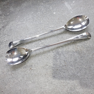 May include: Two shiny, silver serving spoons. One is a solid spoon, and the other has a slotted design. Both have long, slender handles and rounded bowls. The spoons are displayed on a gray, textured surface.