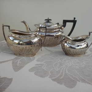 May include: A silver tea set comprising a teapot and two cream jugs, displayed on a floral tablecloth. The teapot features a black handle and lid. The jugs have handles and a decorative, ridged pattern. The set is a classic design.