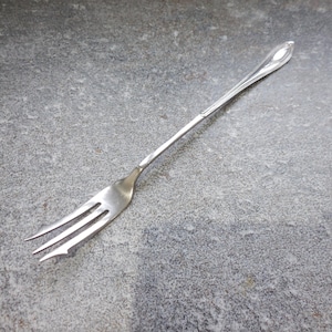 Peut inclure: Une fourchette argentée à trois dents, posée sur une surface grise. La fourchette a un manche décoratif avec une extrémité pointue. Les dents sont légèrement incurvées. Un ustensile pour manger.