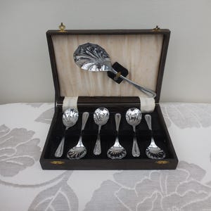 Può includere: Un set di cucchiai da portata argentati e un grande cucchiaio da portata sono esposti in una scatola di legno scuro con interno color crema. I cucchiai presentano decorazioni floreali ornate. La scatola è aperta, rivelando i cucchiai.