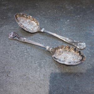 Pode incluir: Duas colheres de prata ornamentadas com padrões florais detalhados. As colheres têm cabos longos e finos e tigelas grandes e ovais. As colheres são exibidas em uma superfície cinza, mostrando seus intrincados desenhos e superfícies reflexivas.