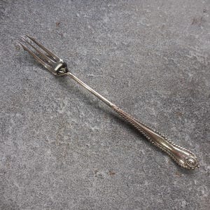 May include: A silver serving fork with three tines, a long, slender handle, and decorative detailing. The handle features a twisted rope design and an oval end. The fork is likely vintage or antique.