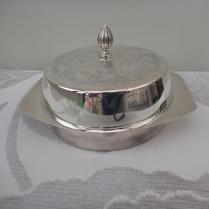 Puede incluir: Una mantequera plateada con tapa abovedada y un adorno decorativo. La tapa y la base tienen una superficie reflectante, y la tapa está adornada con un diseño floral en relieve. El plato se asienta sobre una base cuadrada.