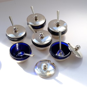 May include: A set of silver salt cellars featuring cobalt blue glass interiors. Each cellar is equipped with a silver lid and a small spoon. The cellars are displayed on a white surface, highlighting their refined aesthetic.
